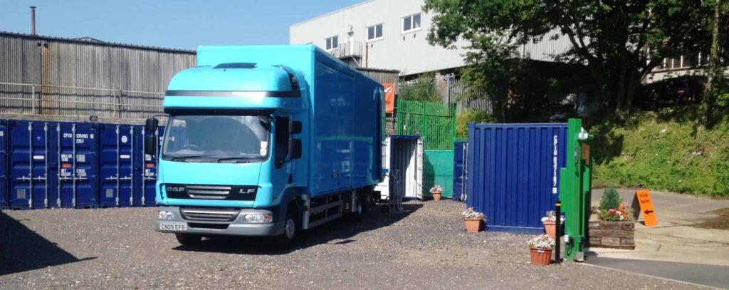 A blue commercial truck parks in a yard surrounded by blue storage containers and potted plants. Industrial buildings and trees are visible in the background, reflecting a busy business storage area. The truck's rear doors are open, and the area is lit by daylight.