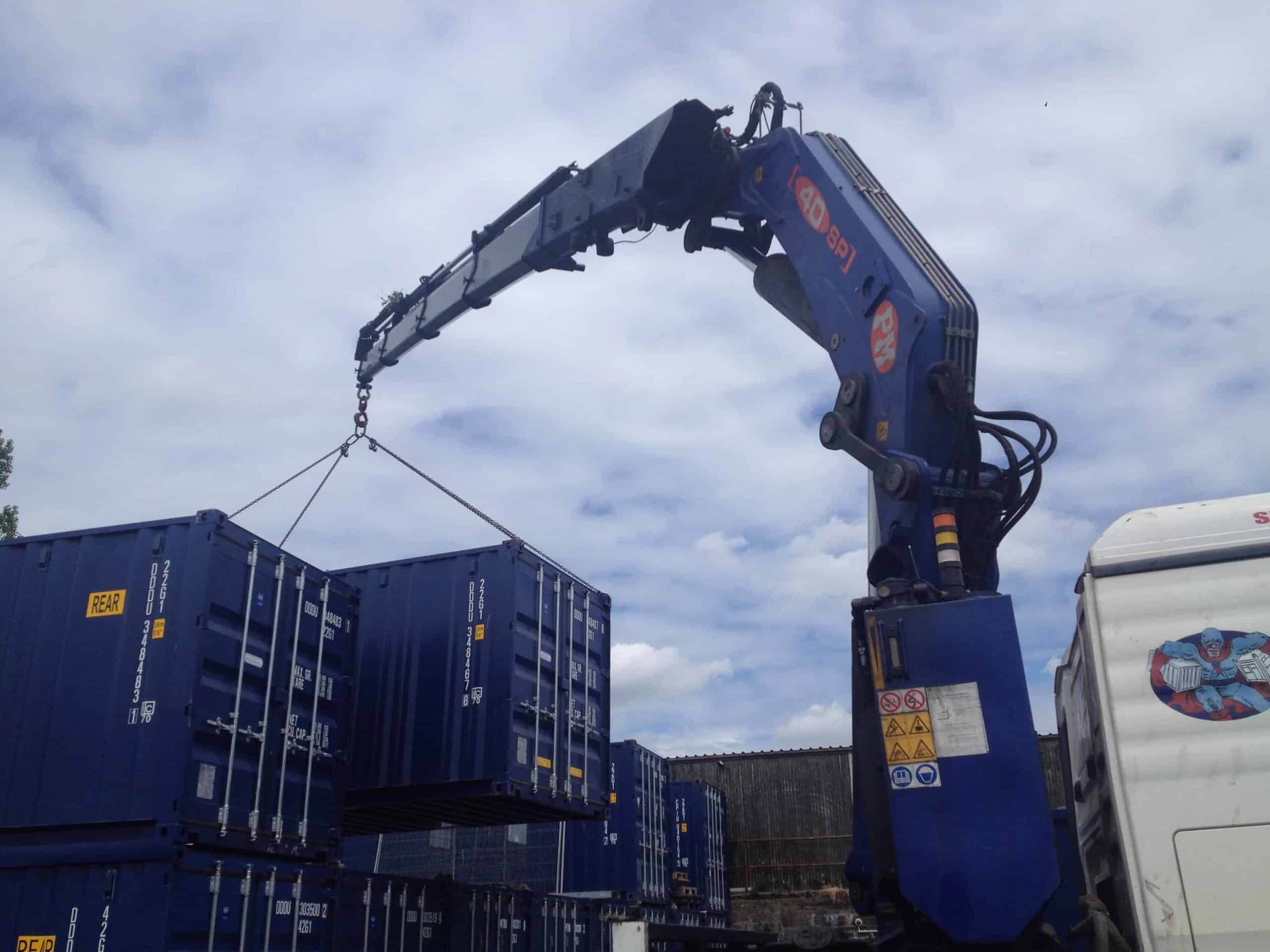 A blue crane mounted on a white truck is lifting a blue shipping container in an industrial yard, where several other self-storage containers are stacked in the background. The sky is partly cloudy.