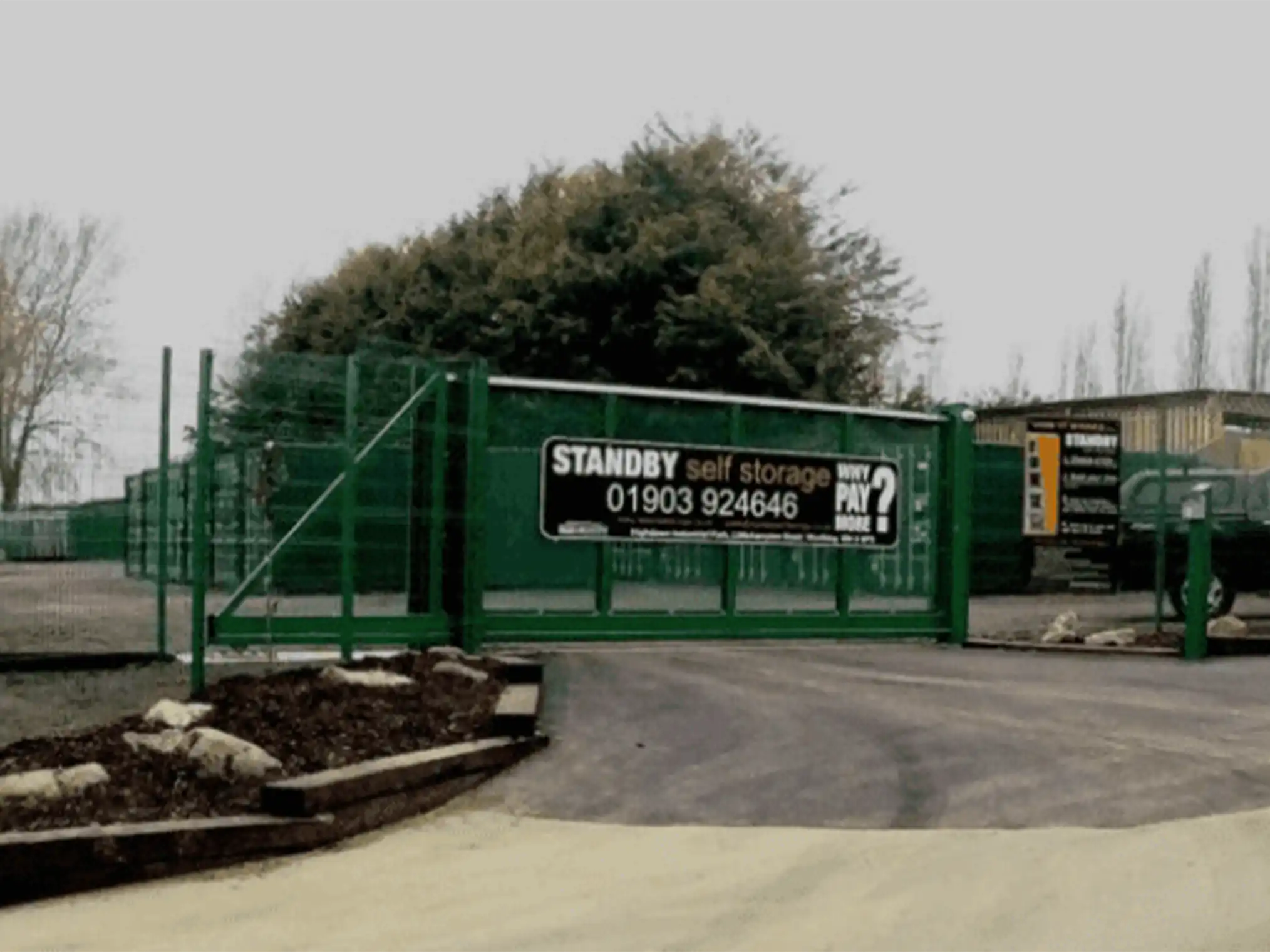 Image shows the entrance to a self-storage facility. A green fence with a closed gate displays a sign reading "STANDBY self storage" with a contact number and "WHY PAY?" in large font. Trees and storage units are visible in the background. The sky is overcast, ideal for personal storage needs.