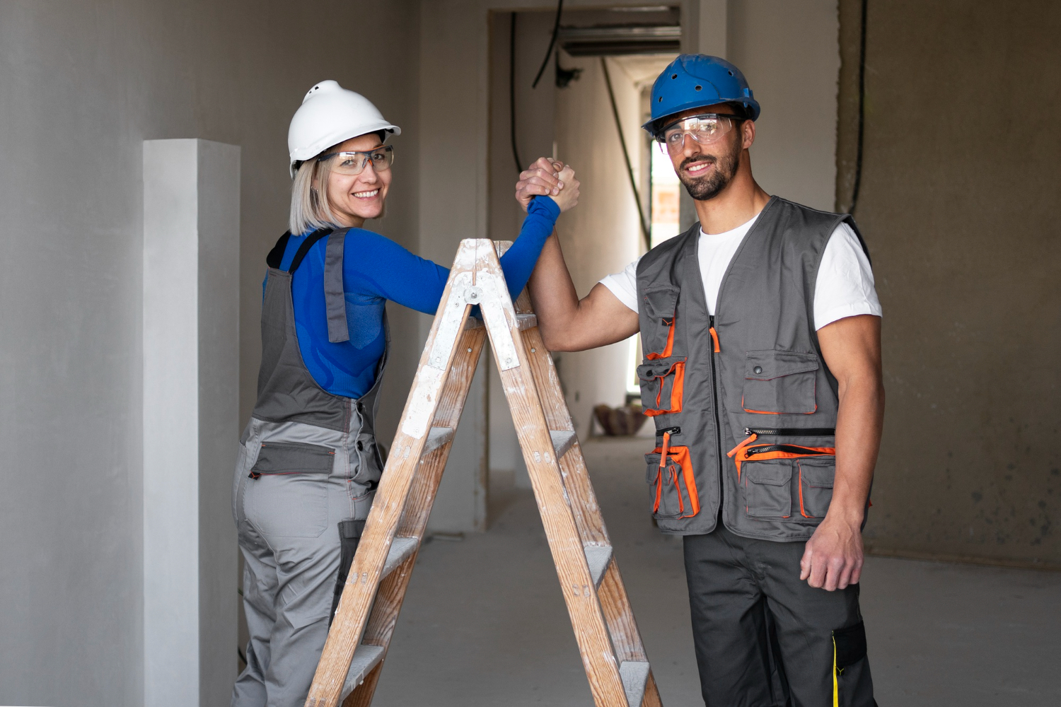 A smiling woman and man in construction gear stand on opposite sides of a wooden ladder inside a building under renovation. Both wear safety helmets and glasses; the woman sports a white helmet, the man a blue one. They hold hands above the ladder, celebrating their progress in the project.