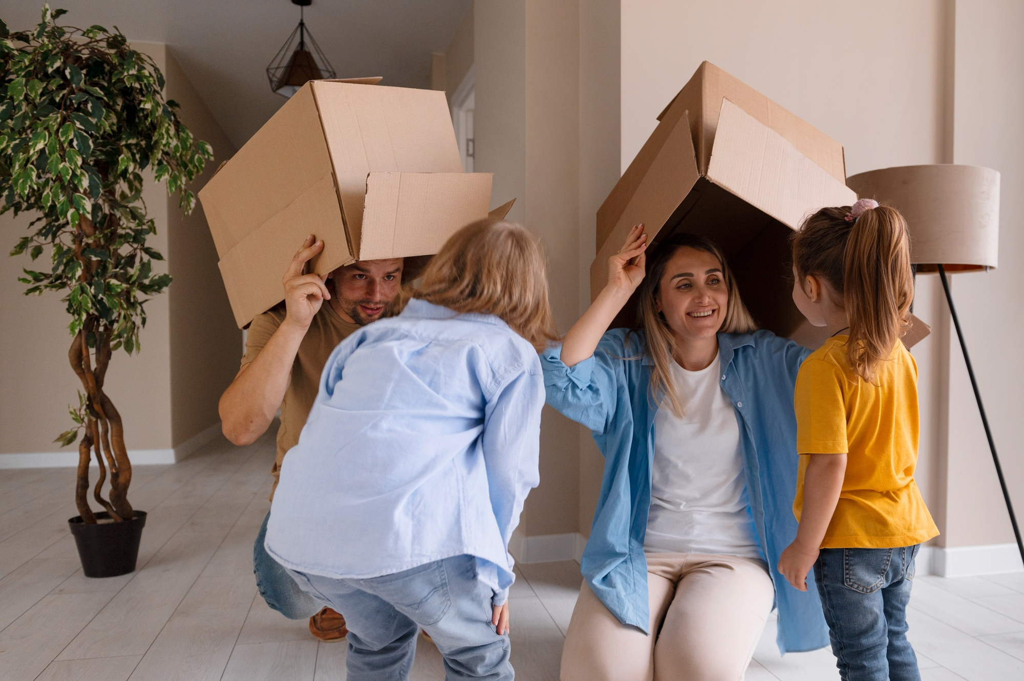 A family of four playfully embraces the chaos of moving house, wearing cardboard boxes on their heads. The parents and two young children smile as they crouch in a living room filled with laughter. Nearby, a tall plant and a lampshade hint at the home's charm amidst the relocating excitement.